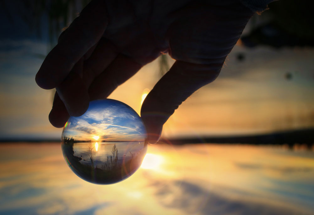 blurred landscape background. in the foreground, a hand is holding a glass sphere. inside the sphere is a clear image of the landscape, which contains a lake and various plants